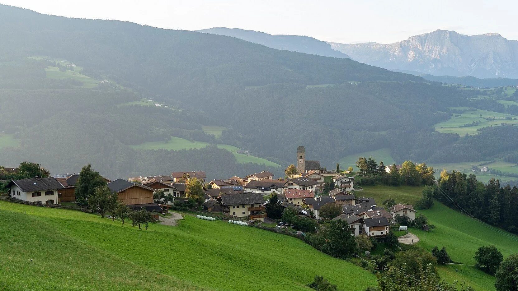 A small village on green rolling hills with the Dolomites beyond